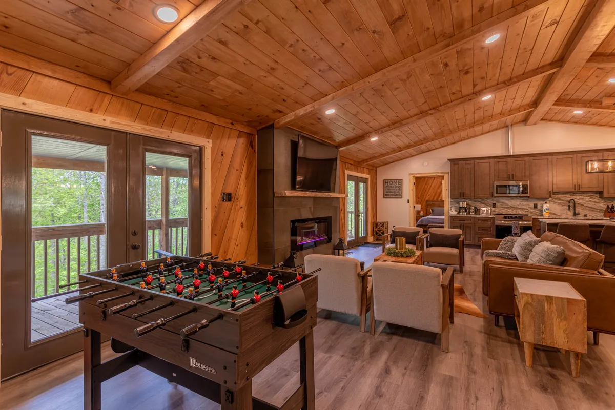 A playful angle on the living space, featuring a foosball table near the seating area, fireplace, and open kitchen. The arrangement emphasizes fun, friendly competition, and the seamless flow between relaxation and entertainment zones.