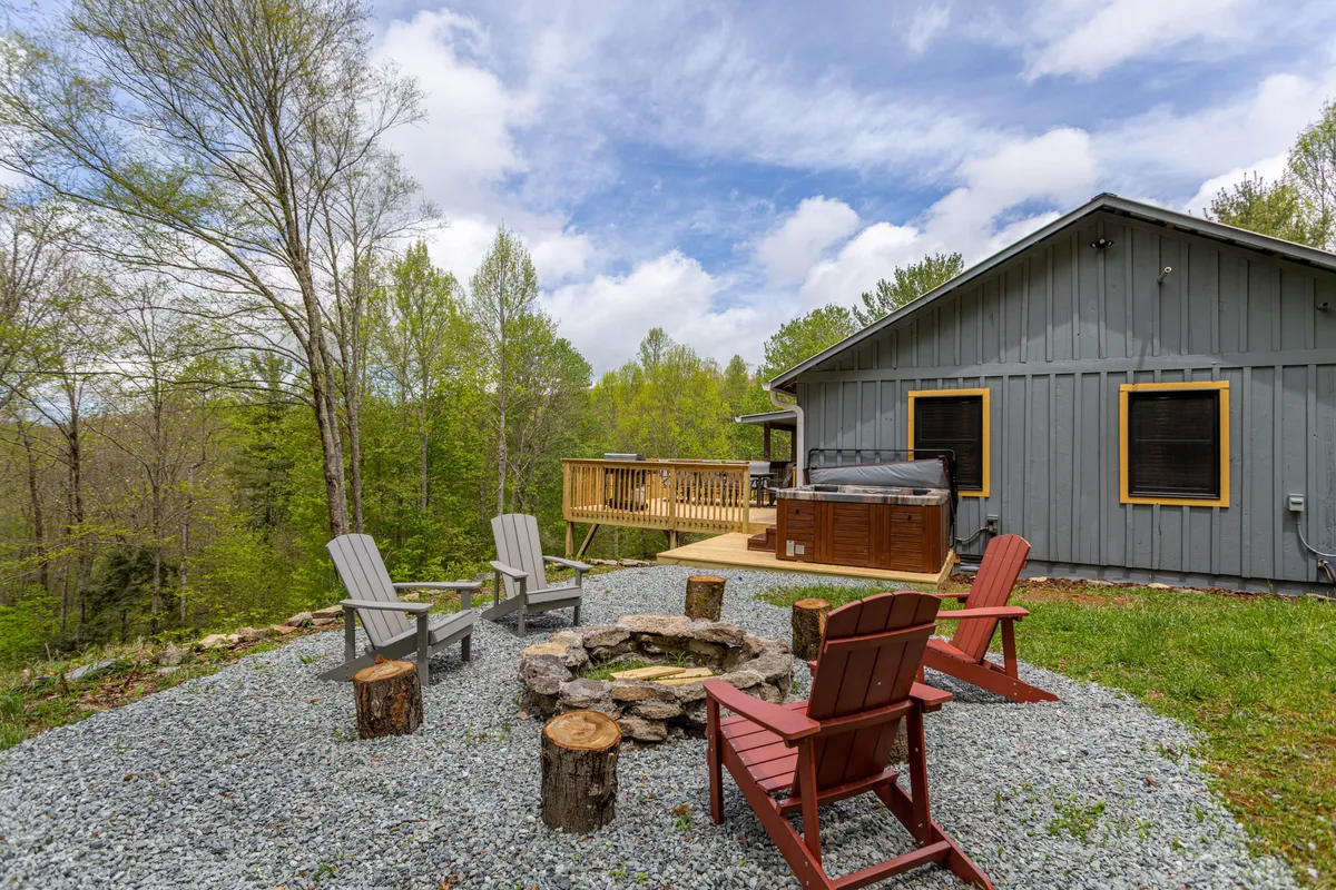 The image displays the exterior of a luxury North Carolina cabin, featuring a spacious deck, a bubbling hot tub, an inviting fire pit surrounded by Adirondack chairs, all set against a backdrop of lush, private woodland. The scene evokes warmth and community, with ample room for relaxation and social gatherings.