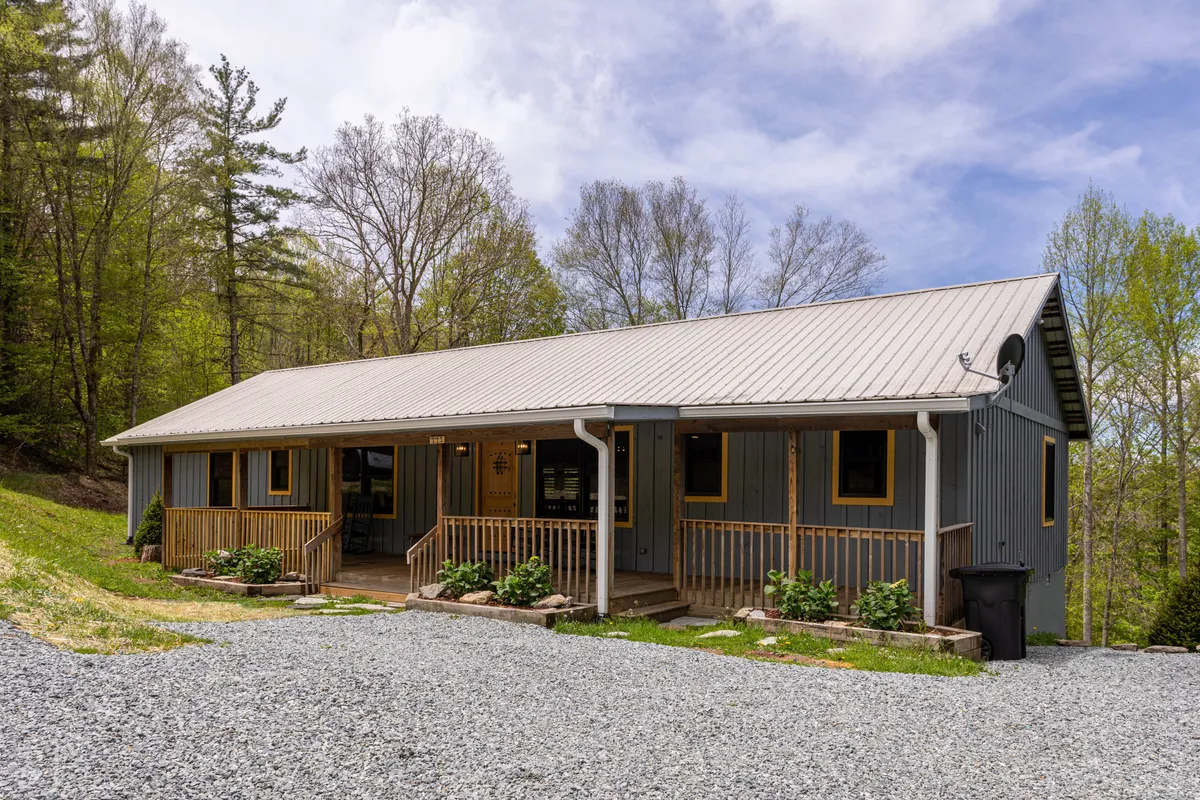 A tranquil exterior view featuring a spacious wooden porch overlooking a gravel driveway, surrounded by lush trees and capped with a metal roof. The inviting front porch is set against a backdrop of peaceful nature, suggesting the perfect spot to unwind after a day of adventure.