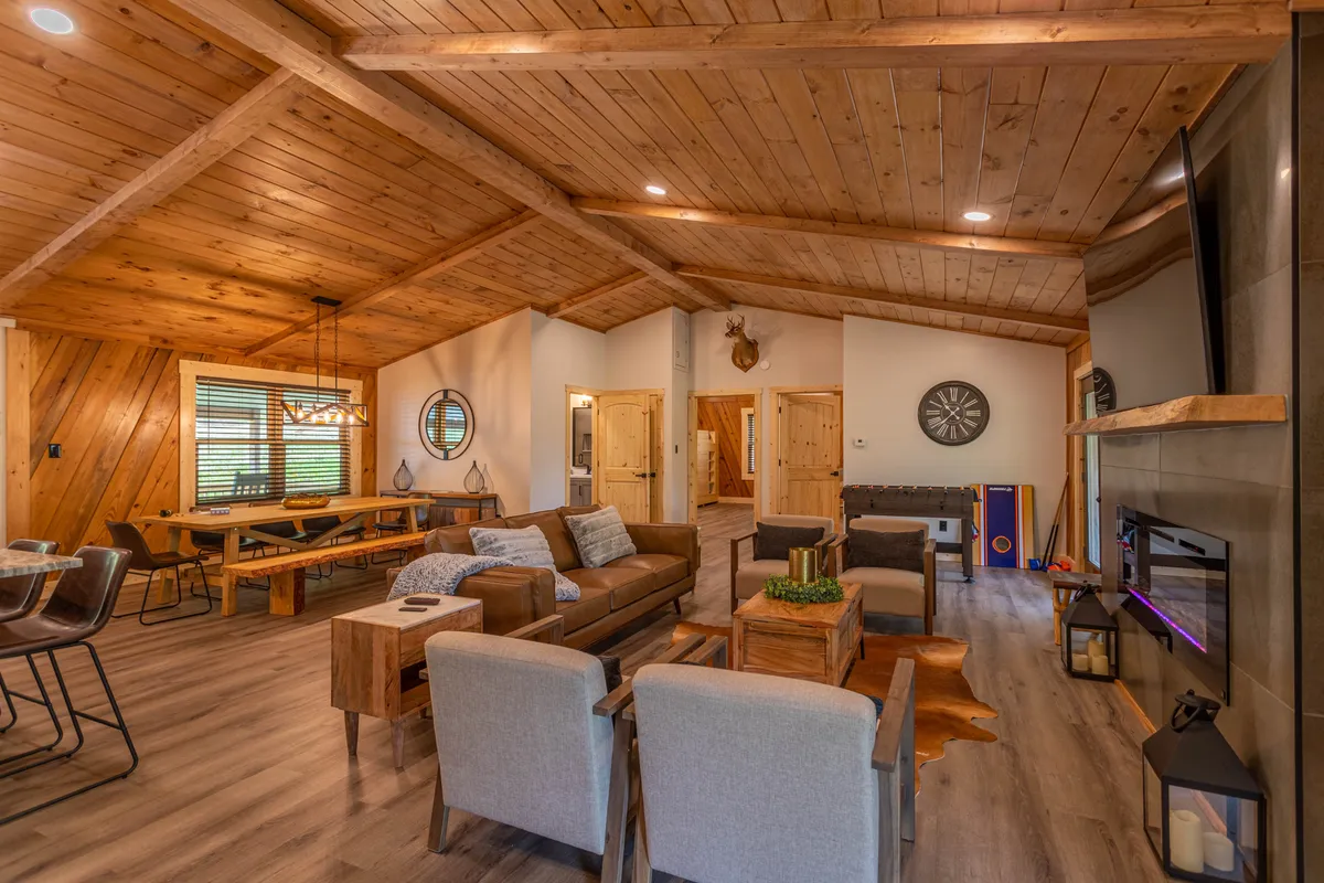 A wide view of the open-concept great room, featuring a wooden ceiling, a large plush sofa, a spacious dining table, and a welcoming fireplace. Decorative wall features add personality while the layout encourages togetherness, with plenty of space for group activities and relaxation.