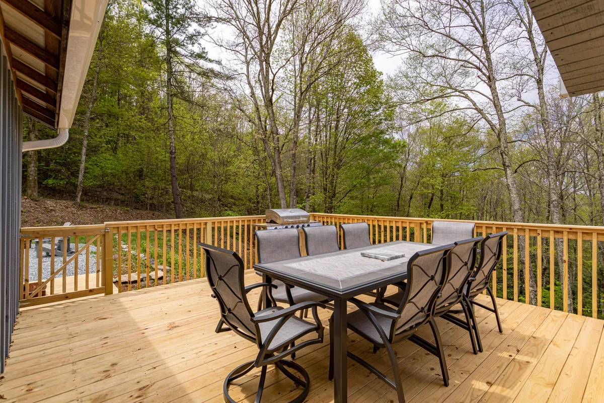 A spacious wooden deck set amidst lush green trees, featuring a dining table with chairs, a gas grill ready for cookouts, and cozy, inviting seating. The surrounding forest offers a sense of privacy and connection to nature, perfect for outdoor gatherings or relaxing with a view.