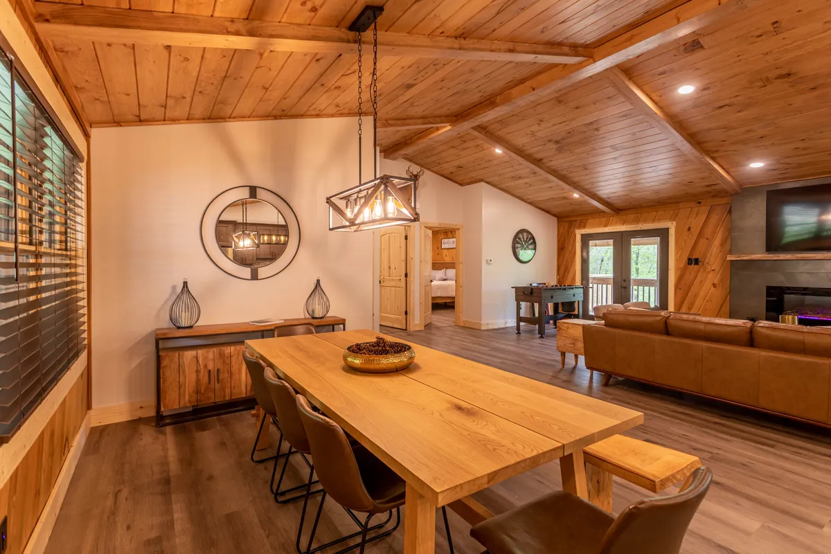 A welcoming dining area with a large table under a wooden ceiling, stylish light fixture, and adjacent seating area. The space is flooded with natural light and designed for shared meals, game nights, and group gatherings.