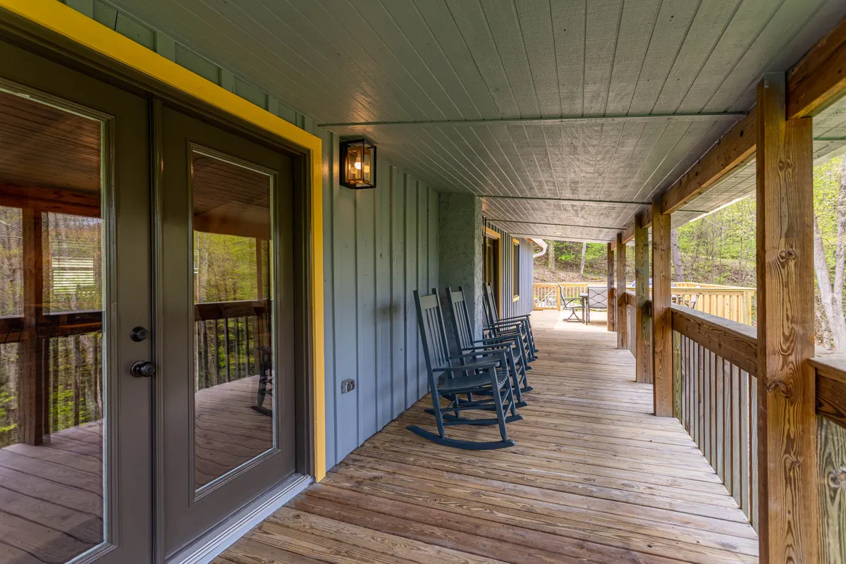 A classic porch scene with wooden rocking chairs and double doors opening to the inside, surrounded by lush greenery. The porch invites relaxation and socializing, with the natural setting just beyond.