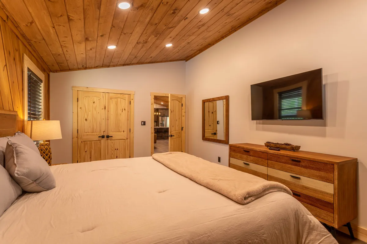 A serene bedroom featuring a king bed with soft, light-colored bedding, a wooden dresser, a TV mounted on the wall, a decorative mirror, and wooden doors. The room is bathed in natural light, creating a peaceful environment for relaxation or movie nights.