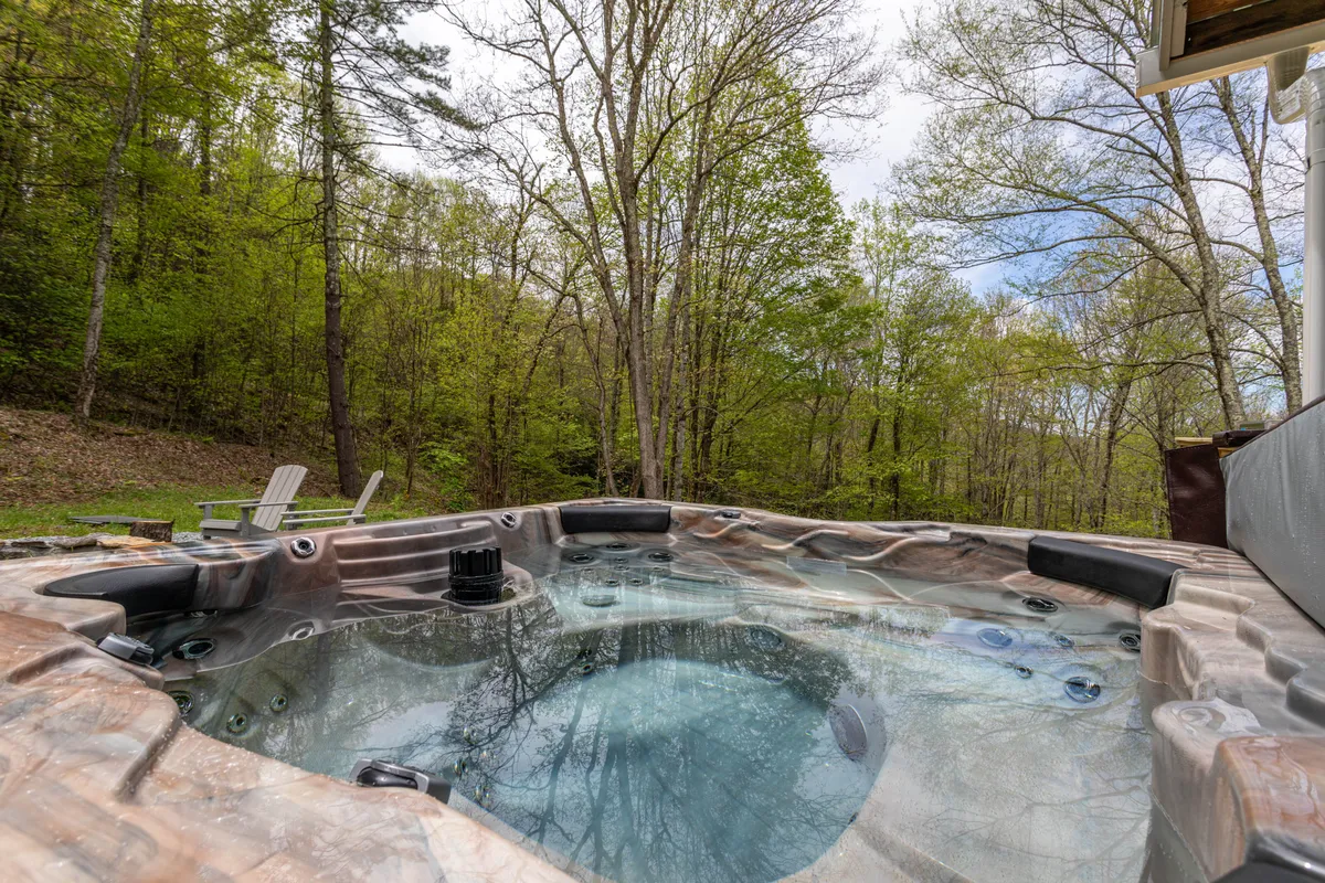 A bubbling hot tub sits on the deck, framed by towering trees and accompanied by lawn chairs for lounging. The area looks serene and private, inviting guests to relax while soaking in natural beauty from the comfort of the hot tub.