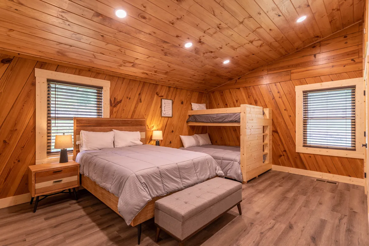A spacious bunk room with a queen bed and a twin-over-queen bunk, all accented by warm wood paneling. The space includes a nightstand and a bench, creating an inviting area ideal for kids or friends to share.