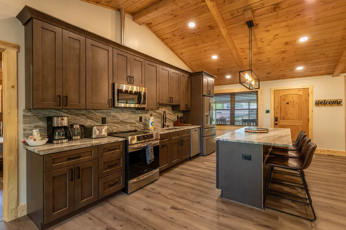 This image shows a bright kitchen with a central island, stainless steel appliances, and wooden cabinets. Natural sunlight streams in through the windows, highlighting the inviting, open layout that connects the kitchen to the outdoors.