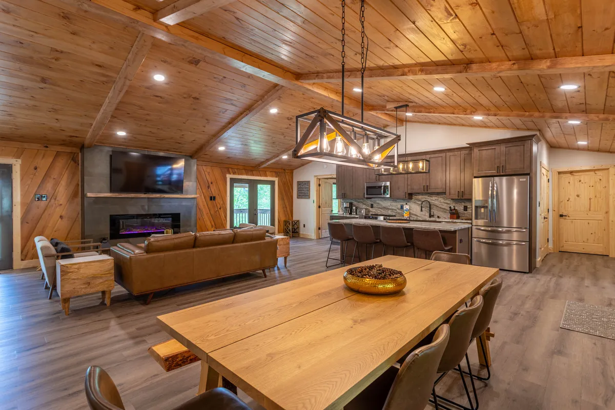 A warm and inviting dining area featuring a dining table, cozy sofa, kitchen island, and a fireplace beneath a chandelier. The open-plan layout is brightened by daylight, providing a welcoming spot for relaxing or gathering after outdoor adventures.